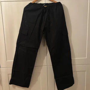 Svarta parachutepants med elastisk midja - Säljer ett par svarta parachutepants i syntetmaterial. De har en loose passform och är perfekta för höst och vår. Byxorna har elastisk midja och praktiska fickor med knappar både fram och bak. Aldrig använt, prislapp kvar. Köpt för 400kr. Xs men kan passa S.