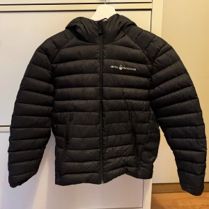 Äkta Sail Raicing JR Spray Down Hood Jacket  - Säljer en svart pufferjacka från Sail Racing i storlek 160. Nästintill nyskick. 80% dun 20% fjäder. Unisex. Från rökfritt och djurfritt hem. Nytvättad och torktumlad. Otroligt skön, snygg och bra passform. Normal i storleken. Nypris 2300:- Jackan har en dragkedja framtill och en praktisk huva. Perfekt för kyliga dagar och har en stilren design med logga på bröstet och huvan. Passar både till vardags och lite mer sportiga aktiviteter.