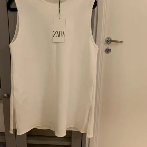 Vit ärmlös topp från Zara - Säljer en stilren vit ärmlös topp från Zara i nytt skick. Perfekt för sommardagar eller som ett basplagg i garderoben. Toppen har en enkel och elegant design som passar till det mesta. Perfekt för både vardag och fest!