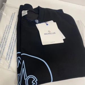Svart tröja från Moncler - Säljer en snygg svart sweatshirt från Moncler i nytt skick. Den har ett stort Moncler-logotryck i blått på framsidan och en liten logga på ärmen. Perfekt för höst och vår, med långa ärmar och ribbade muddar. Superbekväm och stilren, perfekt för både vardag och fest!