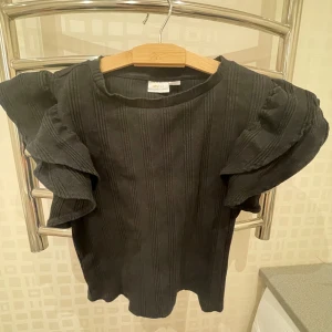 Svart ribbad topp från Gina Tricot - Säljer en snygg svart ribbad topp från Gina Tricot. Den har volangdetaljer på ärmarna som ger en extra touch. Perfekt för både vardag och fest! 🖤
