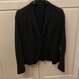 Svart kavaj från Tiger of Sweden - Snygg svart kavaj från Tiger of Sweden i storlek 40. Perfekt för både formella och semi-formella tillfällen. Kavajen har en klassisk design med två knappar framtill och är tillverkad i en blandning av ull och polyester för en bekväm passform. Fickor med lock som kan stoppas in om man tycker de är snyggare - se bild. Ett måste i garderoben för en stilren look! Säljer även ett par tillhörande kostymbyxor, i storlek 38. 
