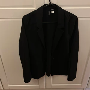 Svart kavaj från H&M - Säljer en stilren svart kavaj från H&M i storlek 38. Rak modell. Ej fodrad. Perfekt för både vardag och fest. Kavajen har en klassisk design med en knapp framtill och långa ärmar. Passar utmärkt till höst- och vårsäsongen. Ett måste i garderoben för en chic look! 🖤