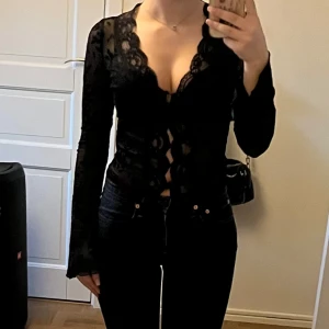 Svart spetsblus med blommönster -  svart spetsblus med blommönster. Blusen har långa ärmar och en djup v-ringning med knytning framtill. Perfekt för en utekväll eller fest! Den är i mycket bra skick och ger en stilren look.