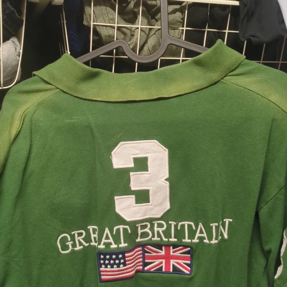 Säljer en grön pikétröja från Ralph Lauren med 'Great Britain' och flaggor broderade på bröstet och ryggen. Tröjan har en klassisk krage och knappar framtill. Perfekt för en avslappnad stil eller en dag på stan. 🏇storlek xxl men sitter som L, solblek på kragen som man ser på bild . Neuletakit & Villapaidat.