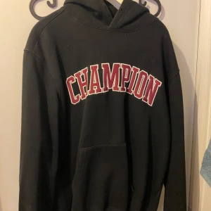 Svart hoodie från Champion - Säljer en svart hoodie från Champion i bra skick. Den har en stor, röd logga på framsidan och en klassisk känguruficka. Perfekt för höst och vinter, och passar både till vardags och träning. Storleken är normal och den är superbekväm att ha på sig. Nypris 1500