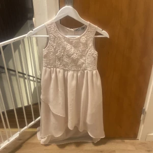 Ljusrosa ärmlös klänning med spetsdetaljer - Söt ljusrosa klänning med spetsdetaljer på överdelen och en flytande kjol. Den är ärmlös och har en dragkedja i ryggen. Perfekt för sommarens fester eller speciella tillfällen. Klänningen har en elegant och romantisk stil.