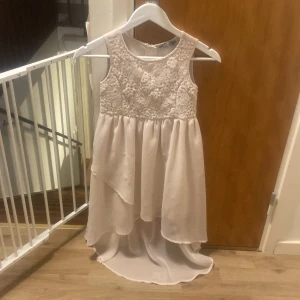 Vit ärmlös klänning med spetsdetaljer - Säljer en superfin vit klänning med spetsdetaljer på överdelen. Den är ärmlös och har en vacker, flytande kjol som är längre bak. Perfekt för sommarens fester eller kanske en midsommarfest. Klänningen har en dragkedja i ryggen för enkel påklädning.