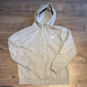 Grå hoodie från Nike - Säljer en grå hoodie från Nike i bra skick. Den har en dragkedja framtill och en praktisk huva med dragsko. Perfekt för höst och vår, eller bara för att chilla hemma. Den är långärmad och gjord i mjukt bomullsmaterial. Passar både för träning och vardag.