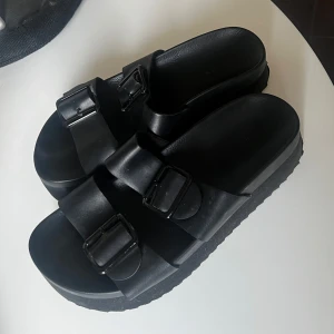 Svarta sandaler med spännen - Säljer ett par svarta sandaler i syntetmaterial med två justerbara spännen. Perfekta för sommaren med en bekväm passform och stilren design. De har en tjock sula som ger extra komfort. Passar både till vardags och avslappnade tillfällen.