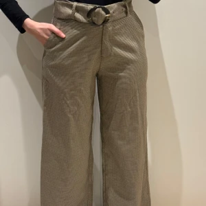 byxa från NA-KD - Säljer ett par snygga rutiga byxor från NA-KD i storlek S. De är högmidjade med ett brett bälte och en cool rund spänne. Byxorna har en loose fit. Materialet känns mycket bekvämt och stilrent.