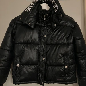 Svart pufferjacka från Calvin Klein - Säljer en snygg svart pufferjacka från Calvin Klein. Den är i mycket bra skick, använt en gång och har en logga på kragen. Jackan har dragkedja och knappar framtill, samt fickor med knappar. Perfekt nu inför vinter. Står storlekn 10 ,passar mig som är xxs/xs! 