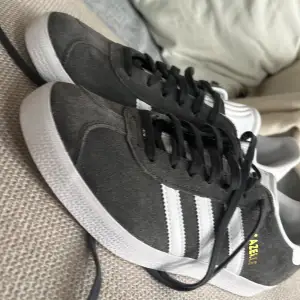 Säljer ett par snygga grå Adidas Gazelle sneakers i mocka med vita detaljer och snörning. De har den klassiska tre-randiga designen och Adidas-loggan på hälen. Perfekta för både vardag och fest! Skorna är i mycket bra skick och kommer med extra vita skosnören.