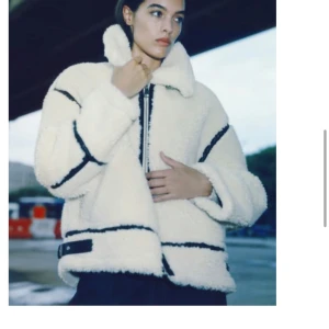 Vit teddyjacka med svarta detaljer - Supermysig vit teddyjacka med svarta detaljer och dragkedja framtill. Jackan har en loose passform och långa ärmar, perfekt för kyliga höst- och vinterdagar. Den har också en cool bältesdetalj vid midjan. Perfekt för att hålla stilen och värmen samtidigt!