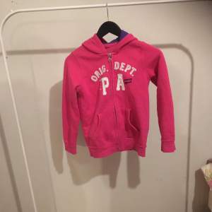 Säljer en snygg rosa hoodie med texten 'Orig. Dept PA' på framsidan. Den har en dragkedja och en mysig luva med lila insida. Perfekt för en avslappnad stil och passar bra till både jeans och leggings. Använd ett par gånger men i mycket bra skick!