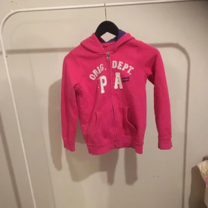 Rosa hoodie med tryck - Säljer en snygg rosa hoodie med texten 'Orig. Dept PA' på framsidan. Den har en dragkedja och en mysig luva med lila insida. Perfekt för en avslappnad stil och passar bra till både jeans och leggings. Använd ett par gånger men i mycket bra skick!