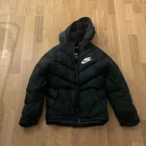Svart pufferjacka från Nike för barn - Säljer en svart pufferjacka från Nike i storlek M. Jackan har en mysig huva och dragkedja framtill. Perfekt för kalla dagar med sin vadderade design. Den är i mycket bra skick och redo för nya äventyr! 🖤