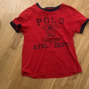 Röd t-shirt från Ralph Lauren - Säljer en snygg röd t-shirt från Ralph Lauren med mörkblå text och detaljer. Den har en klassisk passform och är perfekt för vardagsbruk. Materialet känns mjukt och skönt mot huden. Perfekt för sommardagar eller avslappnade tillfällen. 🏷️
