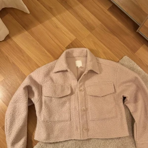 Beige teddyjacka - Säljer en mysig beige teddyjacka med knappar framtill och två stora fickor på bröstet. Jackan har en croppad passform och långa ärmar, perfekt för kyliga dagar. Passar både till jeans och kjol för en trendig look. Helt ny aldrig använd 