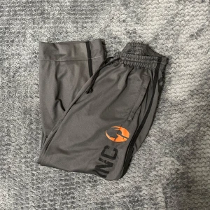 Grå träningsbyxor från GASP - Snygga grå träningsbyxor från GASP med elastisk midja och snörning. Byxorna har en logga i orange och svart på benet och siffran 89 på baksidan. Perfekta för gymmet eller en avslappnad dag. 