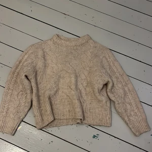 Beige stickad tröja från Gina Tricot - Mysig beige stickad tröja från Gina Tricot med kabelstickat mönster. Tröjan har långa ärmar och en ribbad kant vid halsen, ärmsluten och nederkanten. Perfekt för kyliga dagar!