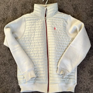 Blå/grön jacka från Polo Ralph Lauren - Säljer en stilren grå jacka från Polo Ralph Lauren med quiltad framsida och röd broderad logga. Jackan har dragkedja och ribbade muddar. Perfekt för kyligare dagar. Även köpt här på Plick som ny så har kvar polo lappen. Skriv vid funderingar!