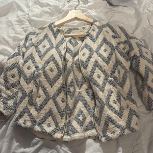Mönstrad jacka i beige och blå - Säljer en unik jacka med geometriskt mönster i beige och blå. Jackan har en normal passform och är långärmad, perfekt för höst och vår. Den är tillverkad i ett mjukt material och har en öppen design utan dragkedja eller knappar.