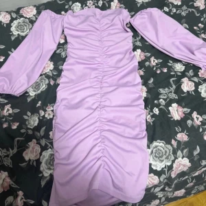 Ljuslila klänning med rynkdetaljer - Säljer en snygg ljuslila klänning med rynkdetaljer längs hela framsidan. Klänningen har långa ärmar och en figurnära passform som framhäver silhuetten. Perfekt för en stilren look.