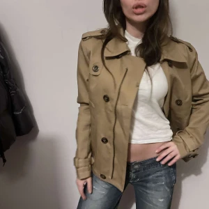 Beige trenchcoat från Black Tag - Snygg beige trenchcoat från Black Tag med knappar och klassisk krage. Perfekt för en stilren look. Jackan har en normal passform och är långärmad.