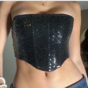 Svart glittrig korsett - Snygg svart korsett med glittriga paljetter och snörning i ryggen. Den är axelbandslös och har en tight passform som framhäver figuren.