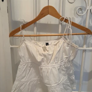 Vit axelbandslös klänning - Söt vit axelbandslös topp med smockdetaljer och volanger. Perfekt för varma sommardagar. Toppen har en luftig passform och är gjord i ett lätt material.