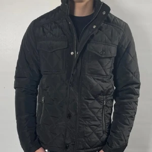 Svart quiltad jacka - Snygg svart quiltad jacka med dragkedja och knappar. Jackan har två bröstfickor med lock och två sidofickor.