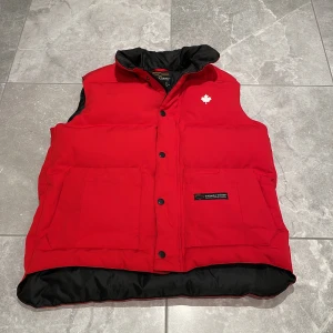 dunväst från Canada Goose - Säljer en dunväst från Canada Goose med svart foder och en broderad vit lönnlöv på bröstet. Västen har knappar och två praktiska fickor framtill. Perfekt för kyliga dagar! Pris kan såklart diskuteras!