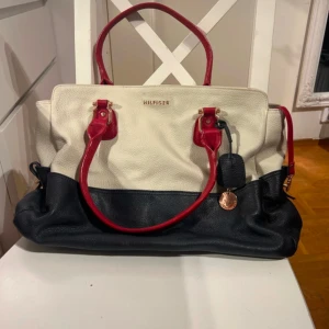 Handväska från Hilfiger ( köpt på BOOZT) - Snygg handväska från Hilfiger i en stilren design. Väskan är i en kombination av vit och svart med röda handtag och detaljer. Den har en praktisk dragkedja och är perfekt för att bära med sig det nödvändigaste. En elegant accessoar för alla tillfällen.köpt på Boozt. 