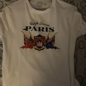 Vit t-shirt från Ralph Lauren - Säljer en vit t-shirt från Ralph Lauren med tryck av texten 'Paris' och flera flaggor. T-shirten har en klassisk rund hals och korta ärmar. Perfekt för en stilren look. Sprillans ny! 