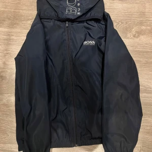 Svart vindjacka från Hugo Boss - Snygg svart vindjacka från Hugo Boss med dragkedja och huva. Jackan har ett stilrent utseende med märkets logga på bröstet. Perfekt för blåsiga dagar.