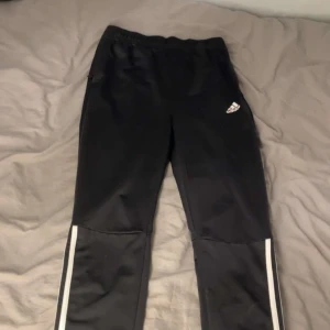 Svarta träningsbyxor från Adidas - Säljer ett par svarta träningsbyxor från Adidas med vita ränder längs sidorna. Byxorna har elastisk midja och är tillverkade i Primegreen-material. Perfekta för sport och träning.