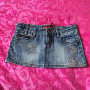 Jeanskjol med broderi - Snygg jeanskjol i kort modell med broderade blommor på framsidan och bakfickorna. Kjolen har en klassisk blå denimfärg och bälteshällor för extra stil. Perfekt för en avslappnad look!