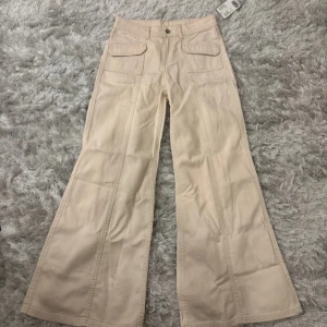 Beige byxor från H&M Divided - Snygga beige byxor från H&M Divided med vida ben och hög midja. Byxorna har framfickor med lock och knappstängning. Perfekta för en trendig look.