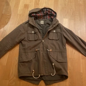 Brun jacka från Brave Soul - Säljer en brun jacka från Brave Soul med rutigt foder och huva. Jackan har knappar och dragsko i midjan för justerbar passform. Perfekt för höst och vår. Jackan har ett bra skick bara att två knappar saknas men 1 finns kvar. 