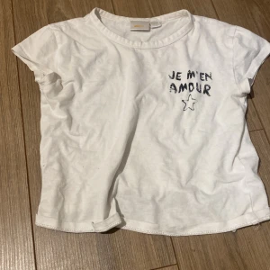 Vit t-shirt från Ginartricot med tryck - Säljer en vit t-shirt från Ginartricot med texten 'JE M'EN AMOUR' och en stjärna tryckt på framsidan. T-shirten har korta ärmar och en avslappnad passform. Perfekt för en casual look!