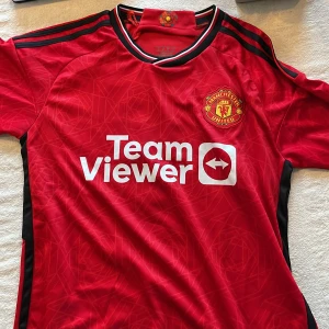 Röd Manchester United fotbollströja +shorts - Säljer en röd Manchester United fotbollströja med korta ärmar. Tröjan har klubbens emblem på bröstet och sponsorlogga 'Team Viewer'. På baksidan finns namnet 'Mount' och nummer 7. Perfekt för fans av laget!