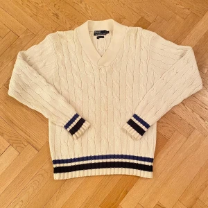 Ralph Lauren Stickad Tröja - Säljer en stilren tjock vit stickad tröja från Ralph Lauren med kabelstickat mönster för att den är för stor för mig. Nypris: 1599 kr. Tröjan har en v-ringad krage och ribbade muddar med blå och mörkblå ränder vid ärmslut och nederkant. Perfekt för en klassisk oldmoney look! Storlek M Hör av er om ni har frågor