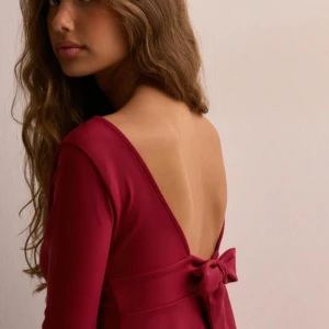 Vinröd topp med öppen rygg och rosett - Elegant vinröd topp med v-ringning och långa ärmar. Toppen har en vacker öppen rygg med en dekorativ rosett. Säljer då den var för liten, helt ny och aldrig använd. Prislappen sitter kvar!