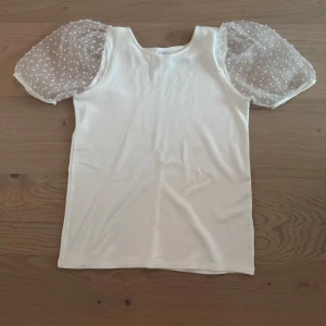 Vit topp med puffärmar - Säljer en söt vit topp från Lindex  med puffiga, transparenta ärmar dekorerade med små vita prickar. Toppen har en rund halsringning och är tillverkad i ett mjukt material. Perfekt för en stilren look!