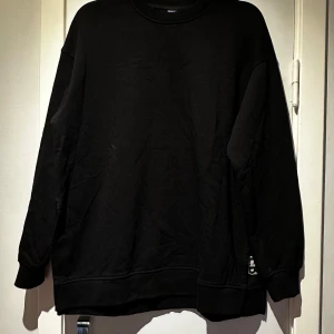 Svart sweatshirt från Stronger - Säljer en svart sweatshirt från Stronger med broderad text på sidan. Tröjan har långa ärmar och en klassisk rund halsringning. Perfekt för en avslappnad stil. Har en slits på båda sidor, se bild (slits  28 cm)   Längd: 69 cm  Brädd: 60 cm Ärmlängd: 51 cm   Nedan kan du se modellen med luva. Notera att detta är modellen utan luva som inte längre säljs.  https://www.strongerlabel.com/se/signature-zinfandel-hoodies-and-sweaters