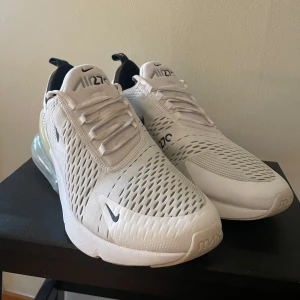 Vita Nike Air Max 270 - Snygga vita Nike Air Max 270 sneakers med en stor synlig luftkudde i hälen för extra komfort. Skorna har en modern design med meshmaterial för bra ventilation och snörning för perfekt passform. Perfekta för en sportig look.