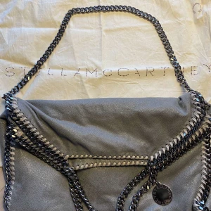 Grå axelväska från Stella McCartney - Snygg grå axelväska från Stella McCartney i mycket bra skick, med kedjedetaljer och en praktisk dragkedja . Dustbag till väskan finns. Skriv om ni vill ha fler bilder på någon detalj eller andra frågor💕