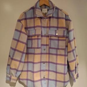 Rutig overshirt från Vila - Säljer en snygg rutig overshirt från Vila i pastellfärger. Jackan har en kombination av beige, lila och blå rutor. Den är långärmad med knappar framtill och två bröstfickor med lock. Perfekt för lager-på-lager-stil!