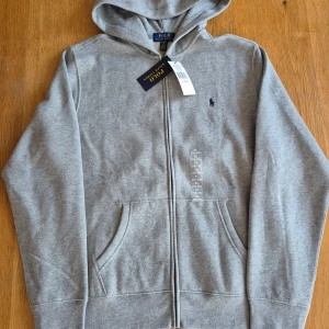 Grå hoodie från Ralph Lauren - Säljer en helt ny hoodie från Ralph Lauren med dragkedja och klassisk logga på bröstet. Lapparna sitter kvar. Sonen råkade få dubletter i födelsedagspresent så nu säljer vi en av dem. 
